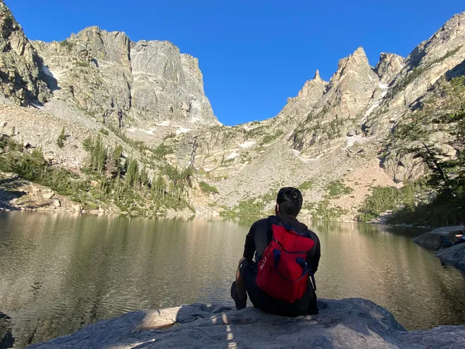 Alok Upadhyay seated at the edge of an alpine lake, looking out toward the mountains